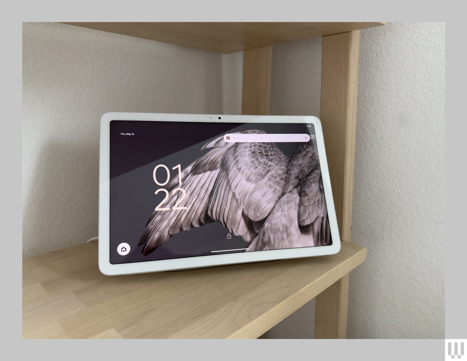 White tablet on light wood shelf with the screen showing the time and a closeup view of bird feathers