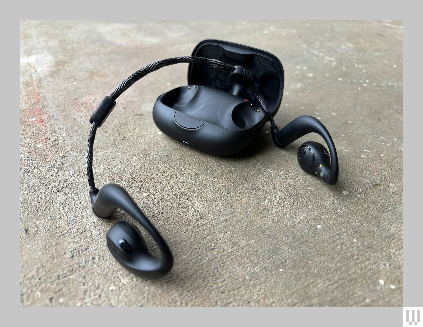 Black rounded rectangular case behind the Anker Soundcore Aerofit Pro headphones which are open and sitting on concrete.