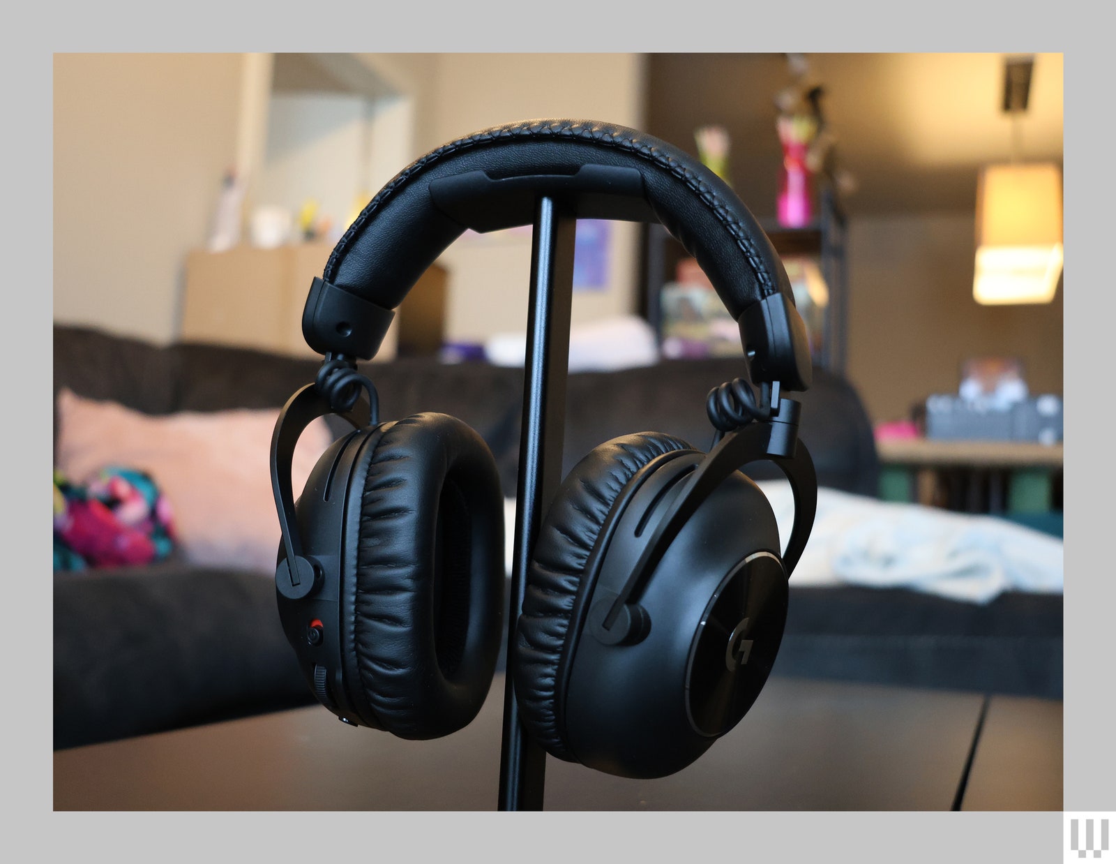 Logitech G Pro headphones on a thin stand. The headphones have a black cushioned band. There is a blue couch and...