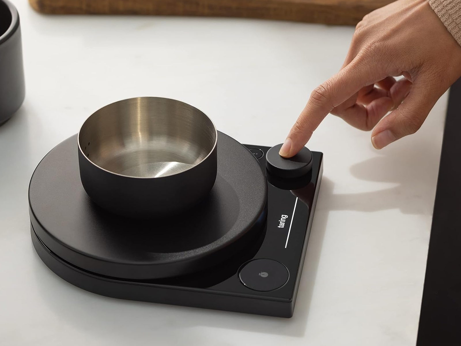 Small black scale with a metal bowl on top sitting on a kitchen counter as a person presses a button on the scale