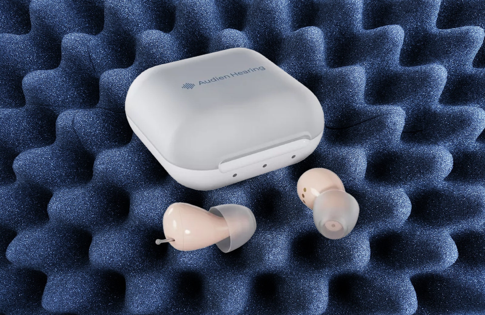 Small rounded closed case beside two beige incanal hearing aides. Image on blue background of soundproof foam.