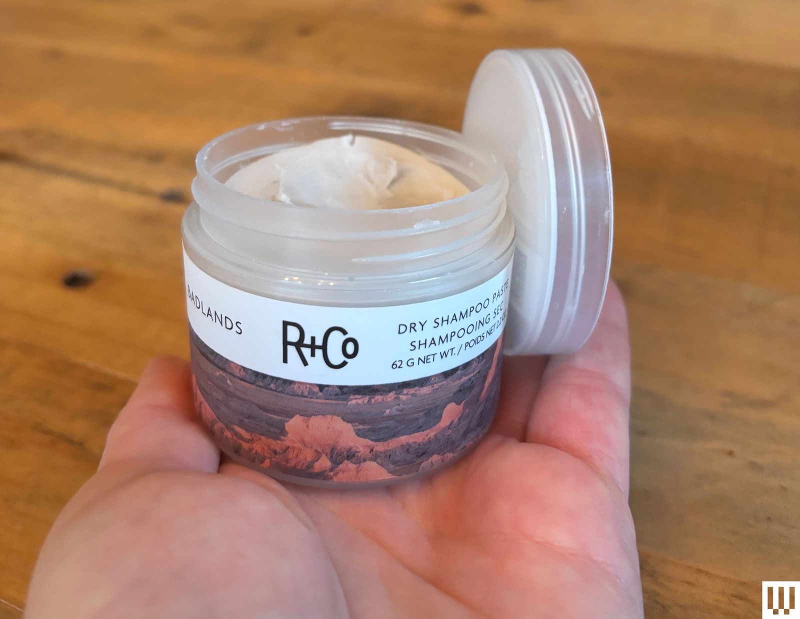 A palm holding R and Co Badlands Dry Shampoo a small jar with beige paste inside and the lid off