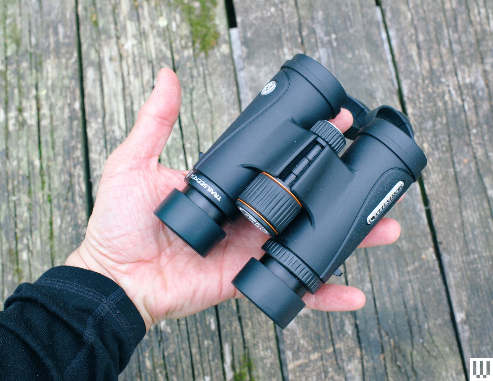 Open palm holding black binoculars neatly folded with a weathered wooden surface in the background.