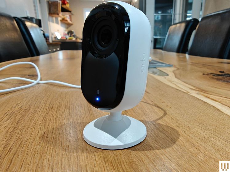 Oval shaped indoor security camera by Arlo with white base and black front, sitting on wooden surface