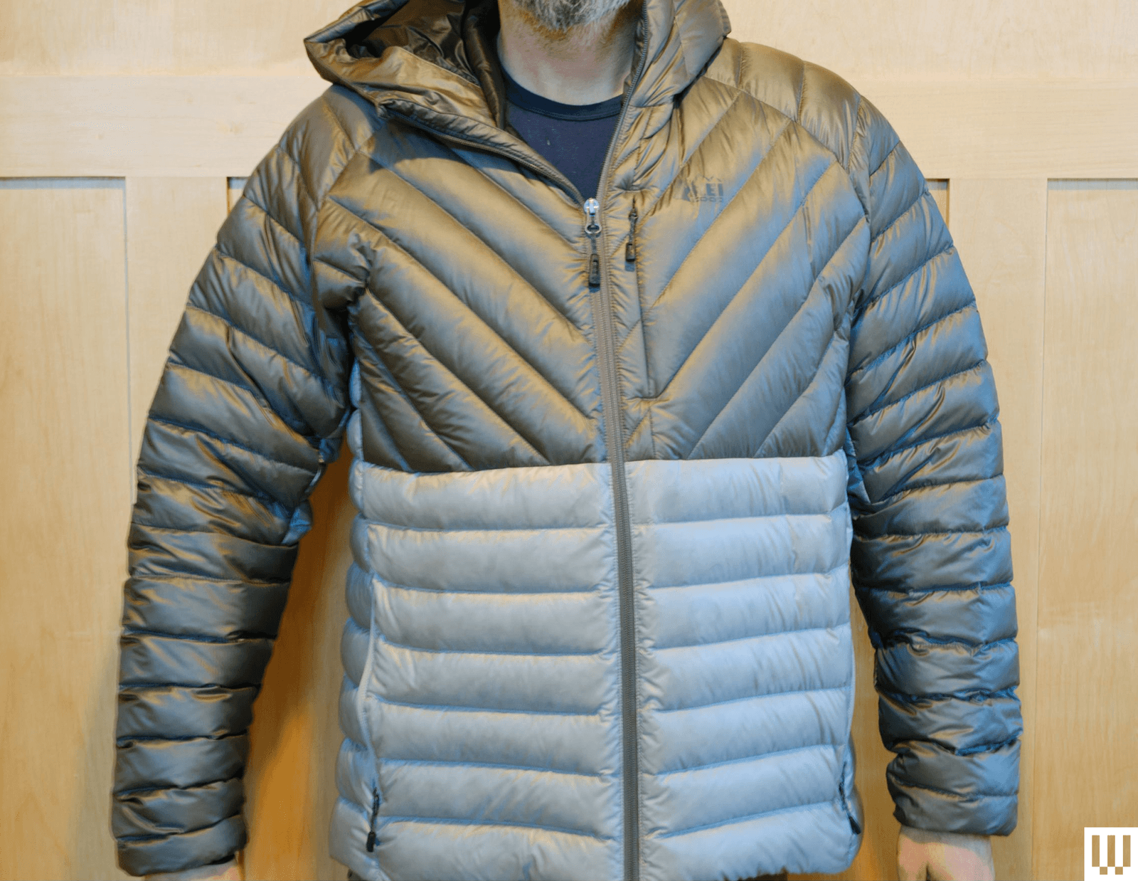 A person with wearing the REI Magma 850 Puffer Jacket in twotone grey standing against a light brown wall
