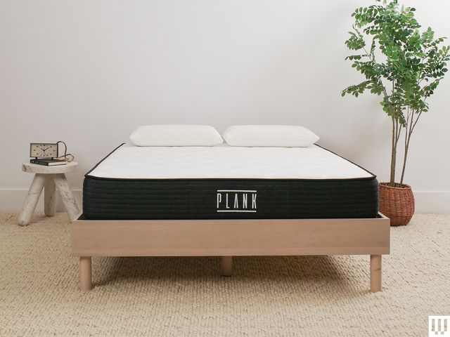 Plank Firm Mattress, a white mattress with blue trim, sitting on a minimalist wooden frame with a nightstand and potted tree on either side