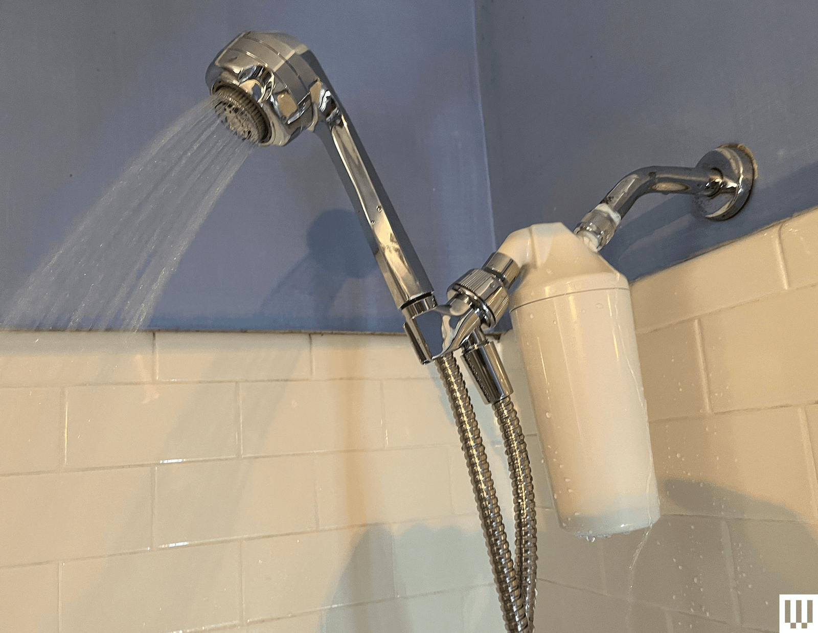 Silver elongated showerhead turned on with white tiles in the background and a white filter attachment connected to the...
