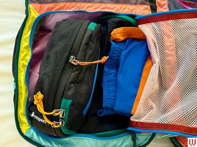 Inside of a travel backpack, showing a mesh pocket unzipped and holding a smaller packing cube and clothing