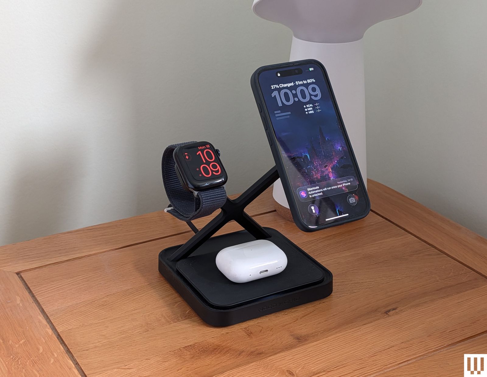 A mobile phone smart watch and case of earbuds on a multipronged stand on top of a wooden nightstand
